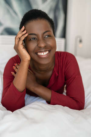 Close up portrait of beautiful Black African woman laying on bed with legs and arms crossed, smiling and looking awayの写真素材