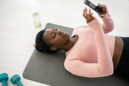 Black African woman laying on yoga mat typing and looking at phone, resting and taking a break from exercising. Shot from above with copy spaceの写真素材