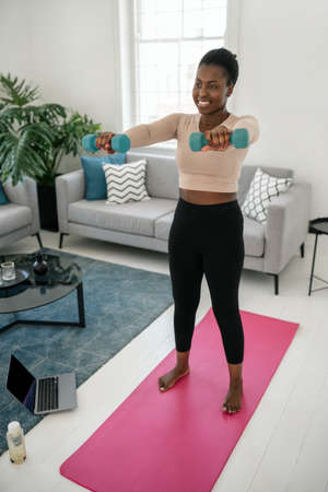 Black African woman exercising following online fitness workout on laptop. Sitting on yoga mat, holding dumbbells in the comfort of modern living room at homeの写真素材