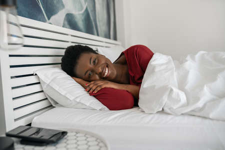 Beautiful African woman laying in bed, looking into camera and smiling, laying comfortably under duvet and on pillows in the morning, just woke upの写真素材