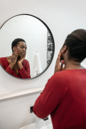 Close up of beautiful Black African woman looking at her face in the bathroom mirror hands on cheeks. Part of her morning self care routineの写真素材