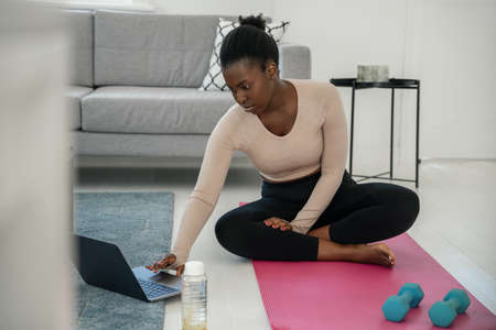 Black African woman following online fitness workout on laptop. Sitting on yoga mat, holding dumbbells in the comfort of modern living room at homeの写真素材