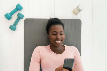 Black African woman laying on yoga mat smiling and looking at phone, resting and taking a break from exercising. Shot from above with copy spaceの写真素材