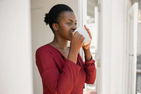 Portrait of a beautiful African woman standing in door way of home, holding a mug, looking into distanceの写真素材