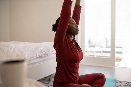Beautiful Black African woman sitting on yoga mat on bedroom floor. Stretching arms upwardsの写真素材