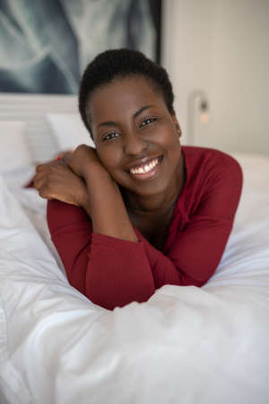 Close up portrait of beautiful Black African woman laying on bed with legs and arms crossed, smiling and looking into the cameraの写真素材