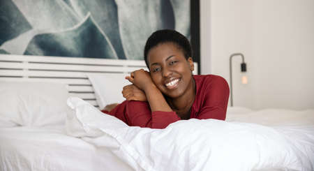 Panoramic portrait of beautiful African woman laying on bed with legs and arms crossed, smiling and looking into the camera with copy spaceの写真素材
