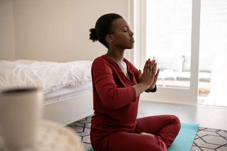Beautiful Black African woman sitting on yoga mat on bedroom floor. Meditating with hands together and eyes closedの写真素材