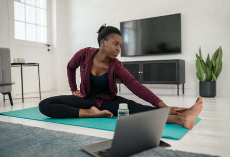 Black African woman following online work out on laptop at home, stretching on yoga mat in living roomの写真素材