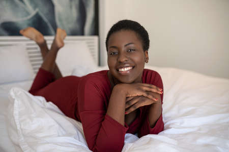 Portrait of beautiful Black African woman laying on bed with legs and arms crossed, smiling and looking into the cameraの写真素材