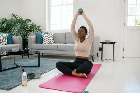 Black African woman following online fitness workout on laptop. Sitting on yoga mat, holding dumbbells in the comfort of modern living room at homeの写真素材
