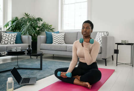 Black African woman following online fitness workout on laptop. Sitting on yoga mat, holding dumbbells in the comfort of modern living room at home.の写真素材