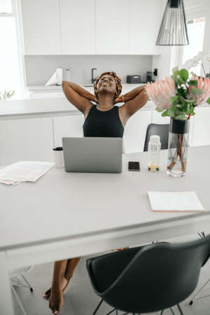 African traditional business woman working from home. Looking up, stretching and arms behind her day dreaming, wearing inspired head scarfの写真素材