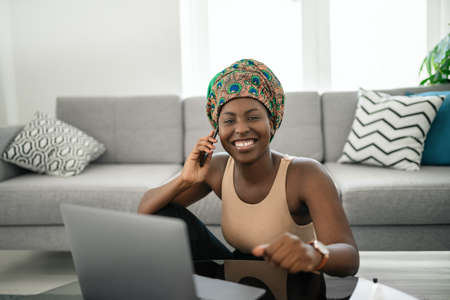 Beautiful young African woman wearing tradition head tie scarf. Sitting at home working on laptop. On phone call smiling and gesturing with handの写真素材