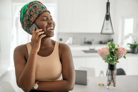 Beautiful African woman at home, smiling and laughing, using mobile smart phone, wearing traditional headscarf and looking into distance with copy spaceの写真素材