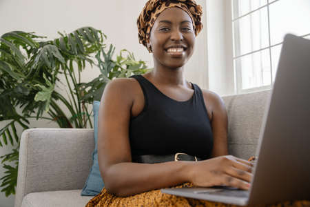 Close up Beautiful traditional young gen z African woman sitting on sofa at home, typing on laptop,...