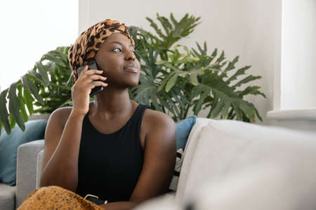 Beautiful young African woman sitting on sofa holding smart phone, looking out of the window, shot with copy spaceの写真素材