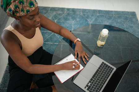 African woman studying from home, looking at laptop, writing down notes. Laptop and smartphone on home office tableの写真素材
