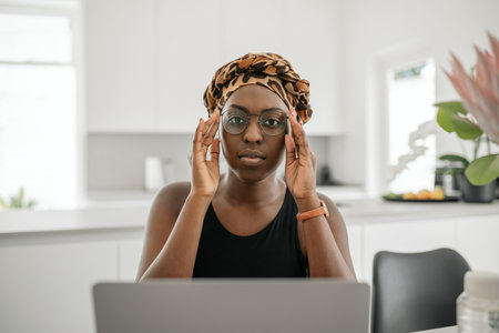 African woman , holding eye glasses. Looking at camera while sitting and working from home. Wearing traditional head scarfの写真素材