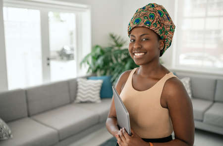 Portrait of beautiful smiling African woman standing in modern home with laptop in hand, wearing a traditional headscarfの写真素材