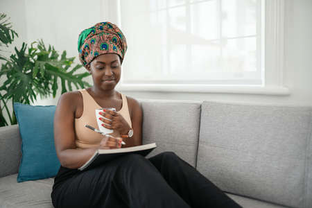 African woman studying from home, writing down notes in book. Relaxing on sofa at homeの写真素材