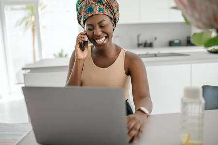 Beautiful young African woman wearing tradition headscarf. Sitting at home working on laptop. On phone call smiling and gesturing with handの写真素材