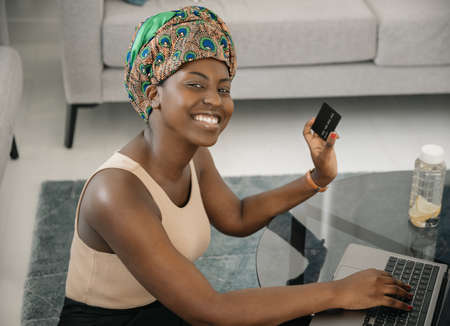 African woman wearing traditional headscarf, making online shopping purchase using laptop and credit card. Sitting by coffee table in lounge at homeの写真素材
