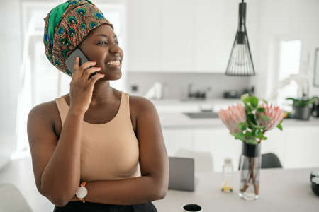 Beautiful African woman at home, smiling, using mobile smart phone, wearing traditional headscarf and looking into distance with copy spaceの写真素材