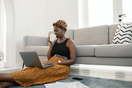 Beautiful African woman sitting on floor in lounge, work meeting on laptop at home with coffee in hand wearing traditional head scarf, sipping on cup of coffeeの写真素材