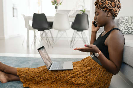 Beautiful African woman sitting on floor in lounge, work call meeting on phone, using laptop at home wearing traditional head tie scarfの写真素材