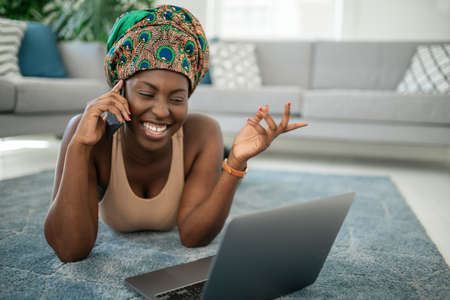Beautiful African woman at home, smiling and laughing, using mobile smart phone, wearing traditional headscarf, surprised and happyの写真素材