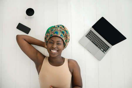 Top view shot of beautiful young African woman laying on white wood flooring, wearing traditional head tie scarf. .With Laptop, mobile phone and cup of coffeeの写真素材