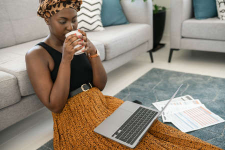 Beautiful African woman sitting on floor in lounge, work meeting on laptop at home with coffee in hand wearing traditional head scarf, sipping on coffeeの写真素材
