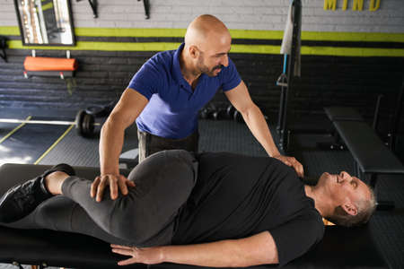 Male personal trainer and physiotherapist assisting mature elderly retired man in stretching....