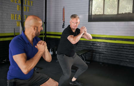 Male personal trainer motivating and assisting elderly senior man while doing squats, dynamic movement exercise in home gym small businessの写真素材