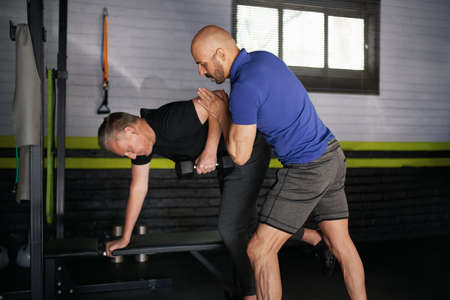 Male personal trainer assisting elderly senior adult man in holding the correct form in doing bench arm rows. Strength and balance training for rehabilitationの写真素材