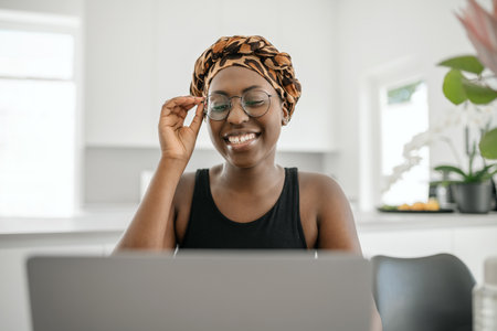 Black African woman smiling, holding eye glasses. Looking at laptop while sitting and working from home. Wearing traditional head scarfの写真素材