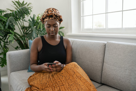 Beautiful traditional young Black gen z African woman sitting on sofa at home, typing on smart phone, smiling, shot with copy spaceの写真素材
