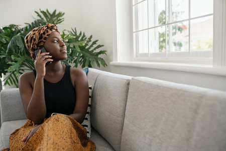 Beautiful young black African woman sitting on sofa holding smart phone, looking out of the window, shot with copy spaceの写真素材