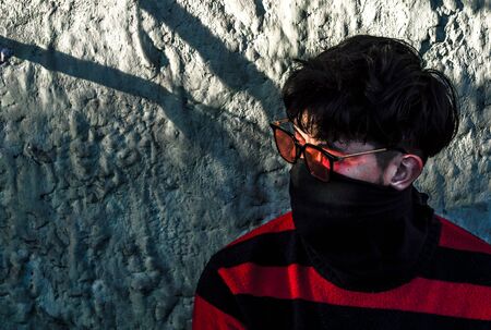 Portrait of a young man with sunglasses and a mask wearing a red striped sweater sitting on the...