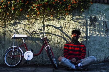 Young Man sitting on the floor with his vintage bicycle in a sunny day in front of a stone wall with plantsの写真素材