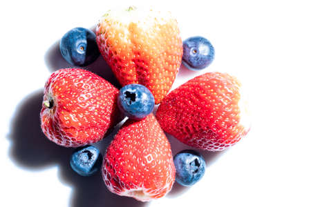 Composition of four very red strawberries forming a cross with several blueberries on the strawberries and around them. Photo on white methacrylate background. Hard illumination with big shadows.の写真素材