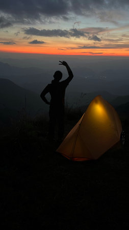 Silhouette of a woman standing in front of a tent on the mountain at sunsetの写真素材