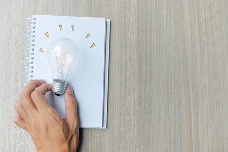 Hand holding light bulb or lamp with notebook on wooden table. New Idea, Creative, Genius and Innovation conceptsの写真素材