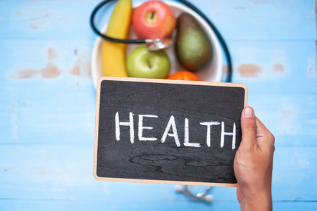hand holding chalkboard over Stethoscope, banana, apple avocado and orange on blue  wooden background. Healthcare and healthy food concept, Top view and Copy space for your textの写真素材