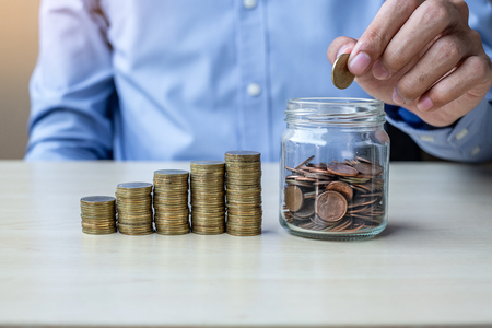 Businessman hand putting golden coin on glass jar with growing money stairs or stack on wooden table. business, investment, retirement planning, finance and Saving for the future conceptsの写真素材