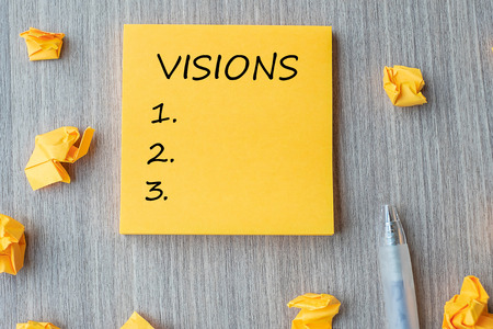 Visions word on yellow note with Businessman holding pen and crumbled paper on wooden table...