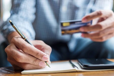 Young casual Business man writing something on notebook while holding credit card and using touchscreen smartphone in the cafe. business, lifestyle, technology, ecommerce and online payment conceptの写真素材
