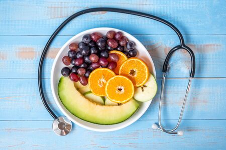 Top view black grapes, red grapes, Valencia orange and melon fruits in white dish with Cardiology Stethoscope on blue wood table. dieting healthy food and healthcare conceptの写真素材