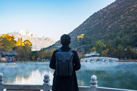 Young man traveler traveling at Black Dragon Pool with Jade Dragon Snow Mountain background, landmark and popular spot for tourists attractions near Lijiang Old Town. Lijiang, Yunnan, Chinaの写真素材